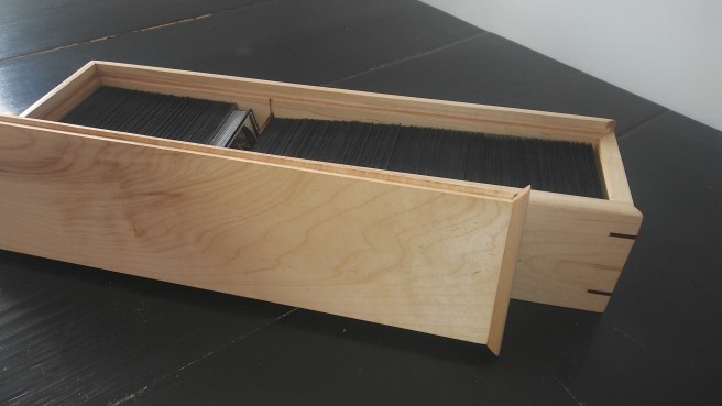 A wooden box sits on a black table. This is known as a "cube" and is used to store cards for the game Magic: the Gathering for the purposes of a repeatable draftable experience.